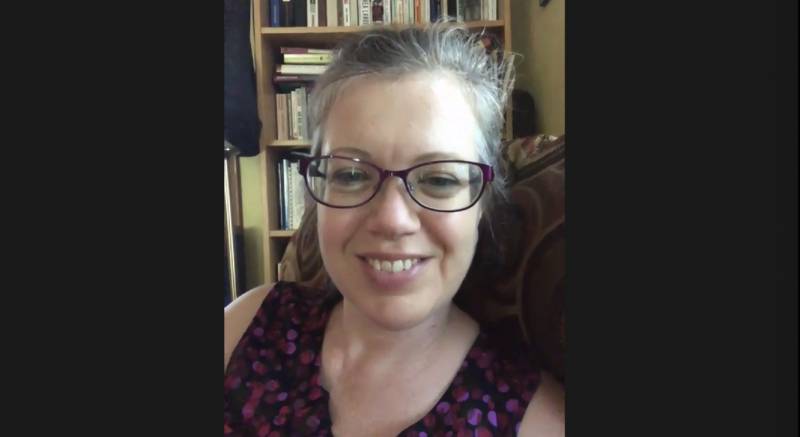 A screenshot of a white woman with her hair pulled back and glasses. She is wearing a sleeveless shirt with purple designs, and there is a bookshelf in the background. Screenshot by Cope Cumpston.
