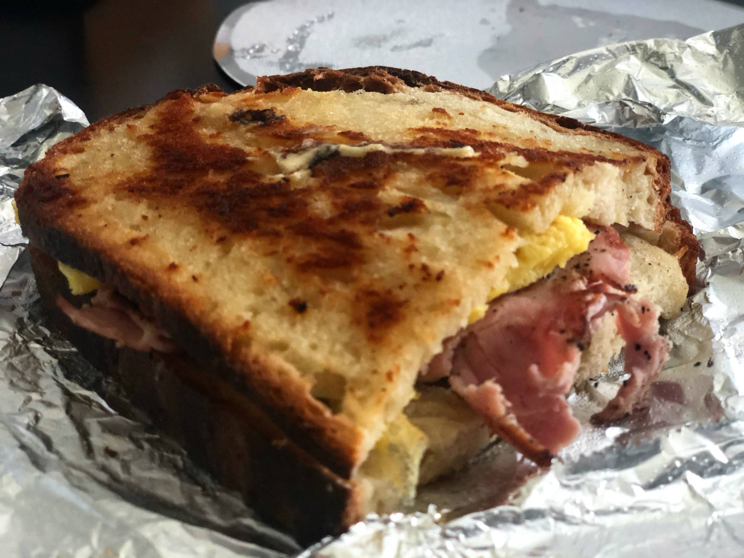 A half sandwich sits on a tin foil wrapper. The bread on the sandwich is toasted and darkened in spots with a bit of ham peeking out of the bread. Photo by Alyssa Buckley.