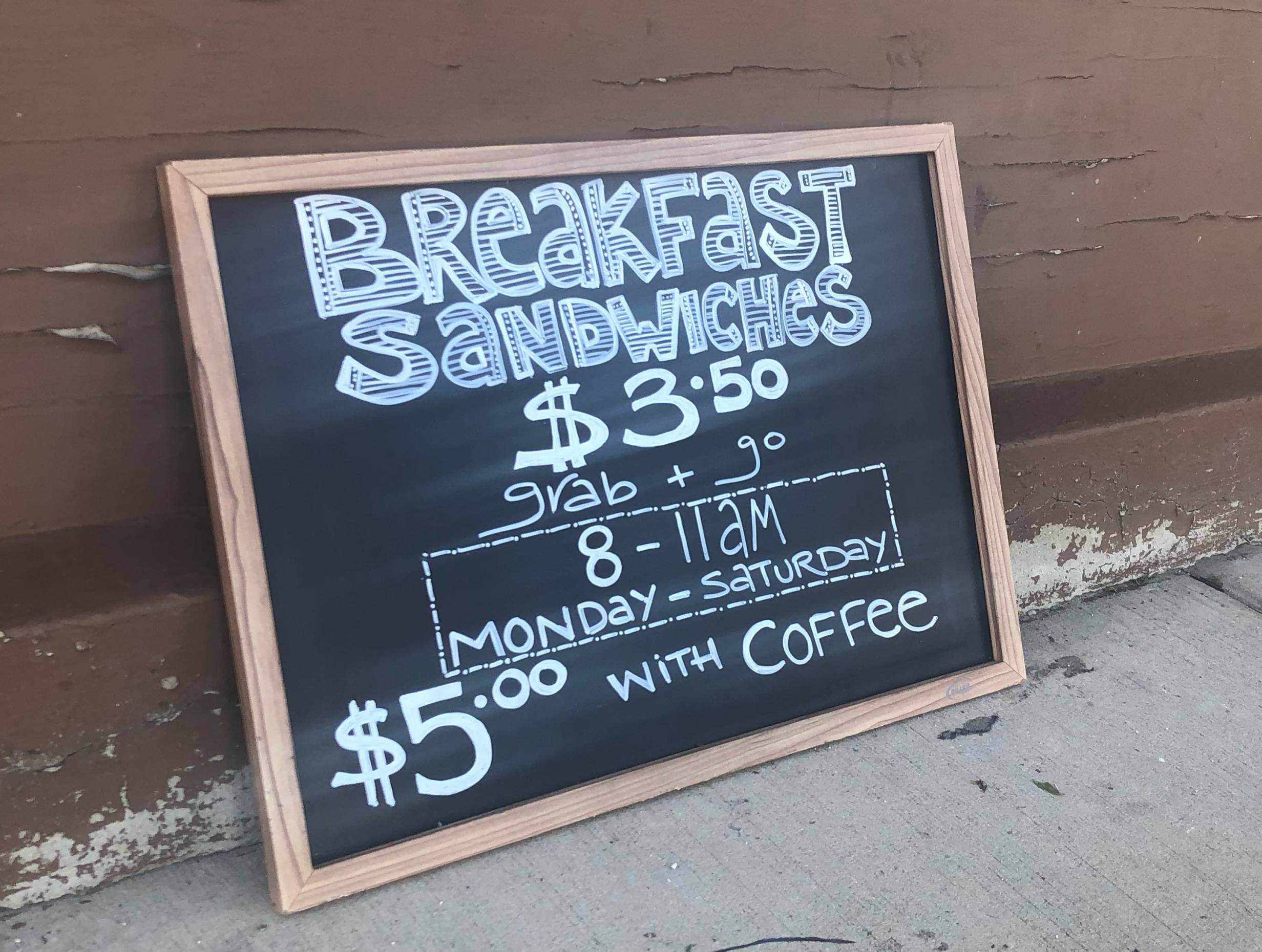 A chalkboard reads: Breakfast Sandwiches with details on hours and pricing. It is leaning against a burgundy building, resting on the sidewalk. Photo by Alyssa Buckley.