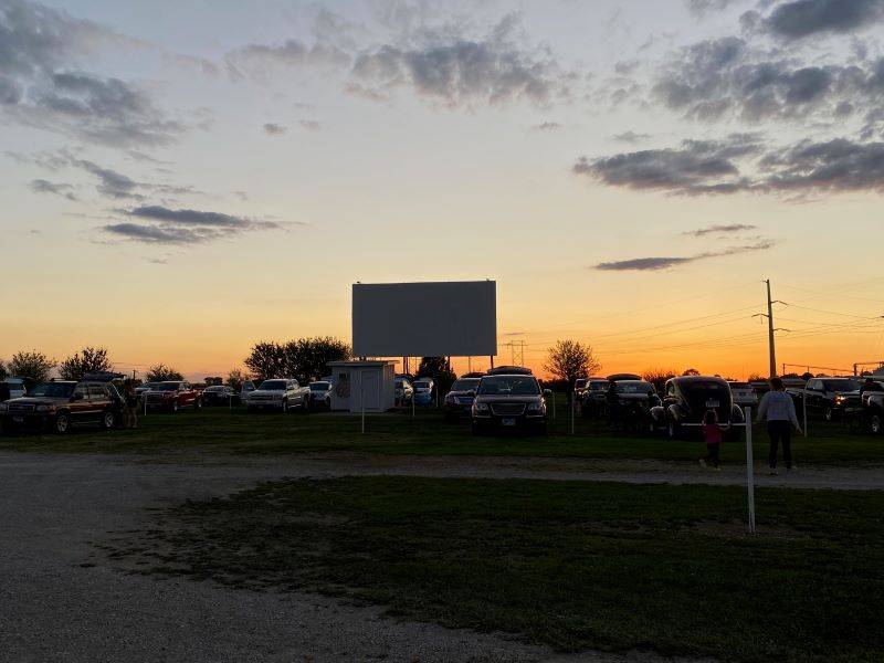 A drive in movie screen with a sunset in the background. Several cars are lined up in front of it. Photo by Julie McClure.
