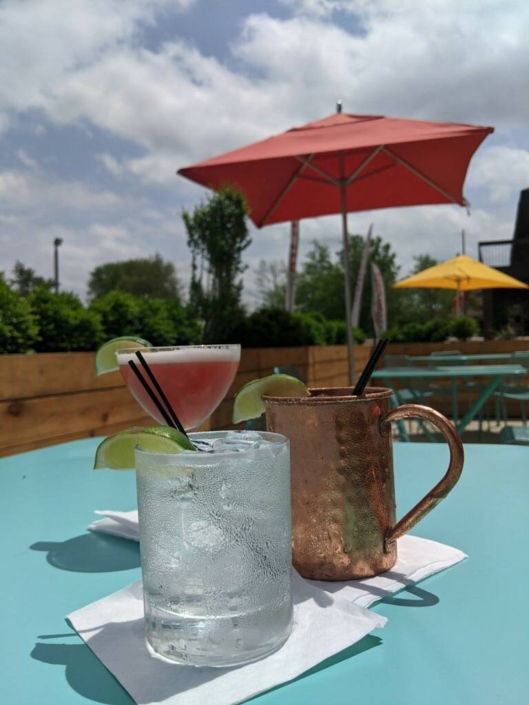  Three drinks sit on a blue table outside of Lodgic. One glass is a tall glass with a pink drink and lime wedge. One is a mule glass with a lime wedge. In the forefront, the third is a glass with a clear liquid and a lime wedge. Photo by Betsy Waller.