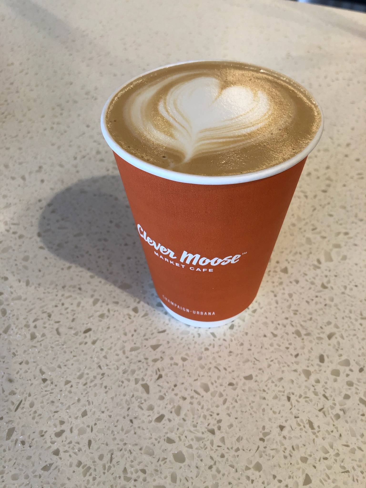 A heart appears Clever Moose's latte which is in an orange paper to go go on a speckled counter. Photo by Betsy Waller.