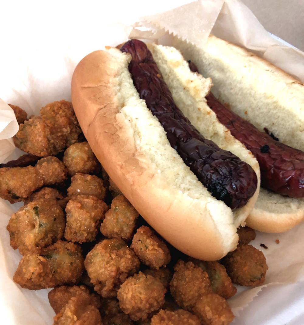 Two grilled sausages in hot dog buns sit on top of pieces of fried okra. The meal is served in a Styrofoam take out containter. Photo by Jessica Hammie. 