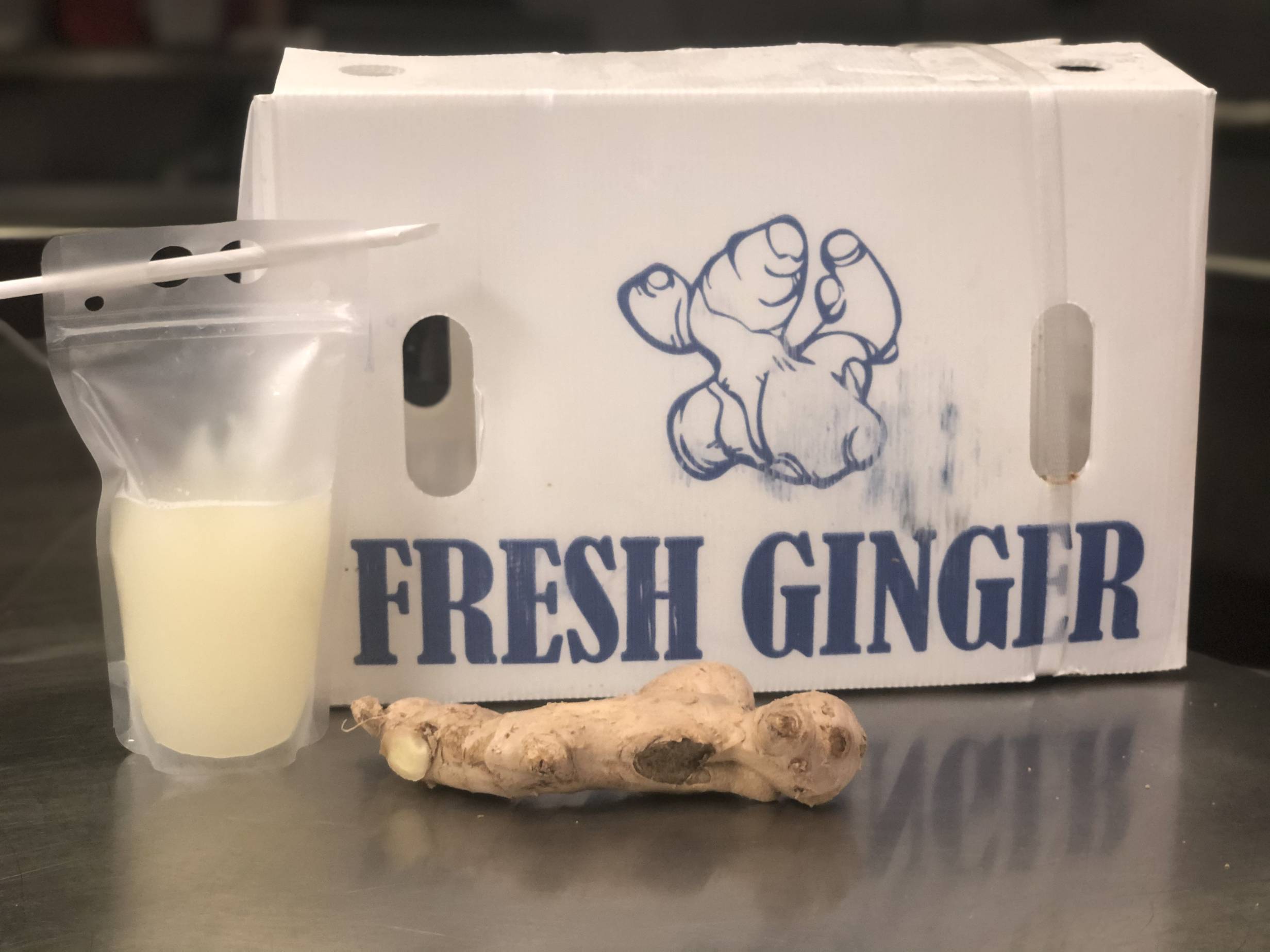 A plastic bag of ginger juice with a paper straw sits on a metal counter with a ginger root in front of a large white box reading 