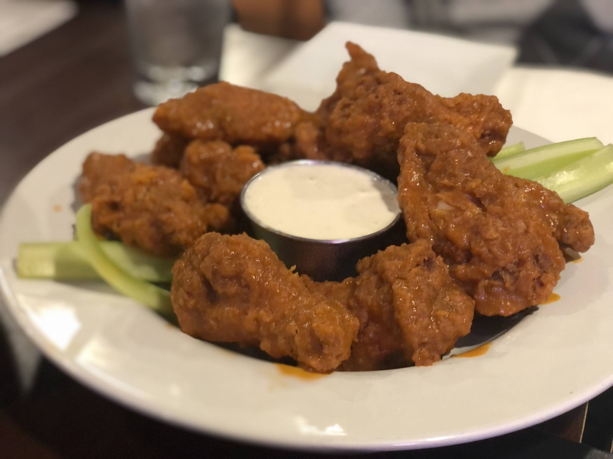 Twelve bone-in fried chicken wings are covered in an orange buffalo sauce on a white plate with thin celery slices and a cup of ranch dressing in the center. Photo by Alyssa Buckley.