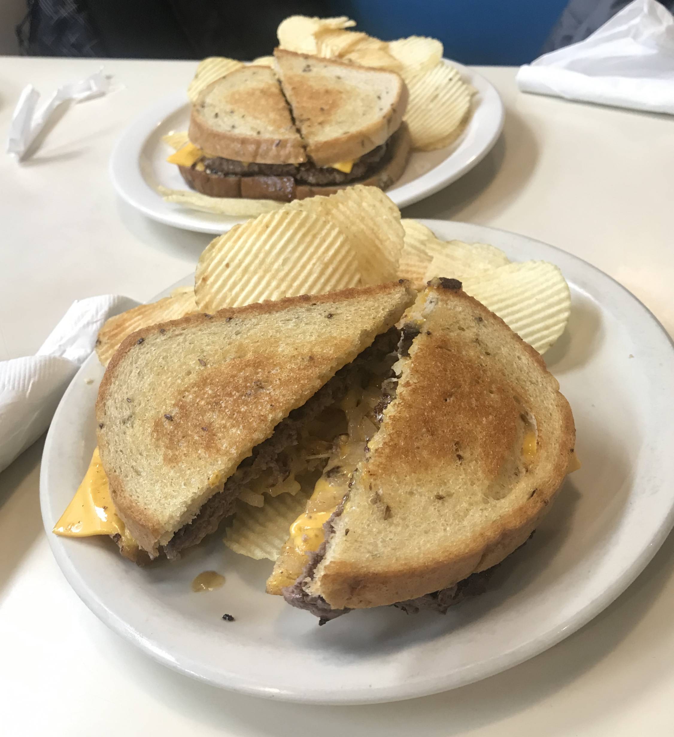 Two patty melts at Merry Annâ€™s served over a bed of potato chips. Both burgers are cut in half and inside the cheese seeps over the side of the buttered and toasted rye bread and a peak of a beef patty and slivers of onion. The plates sit on a white table with utensils and napkins. Photo by Kanea Hughes.
