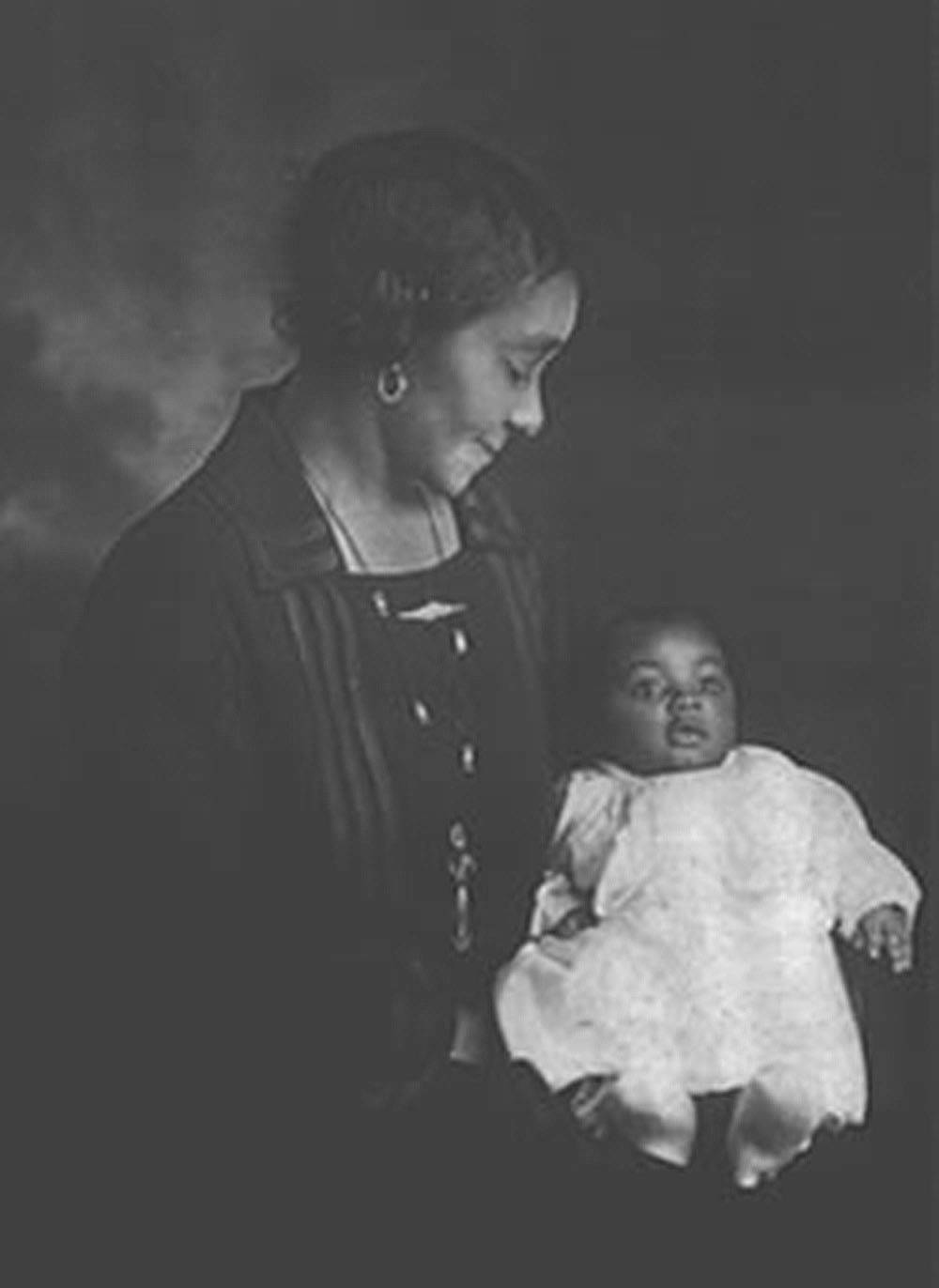 A Black woman holds a Black baby wearing a white garment. The background of the image is dark. Photo provided by Barb Garvey.