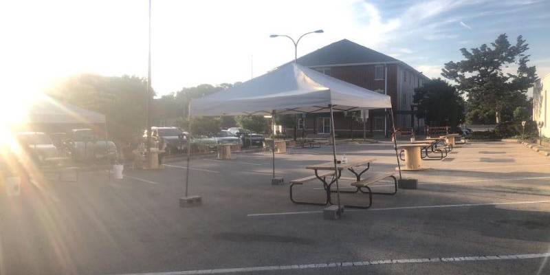 IMAGE: Photo of a parking lot with a sun flare from the left hand side. There's a white top tent in the middle with tables and chairs around it.