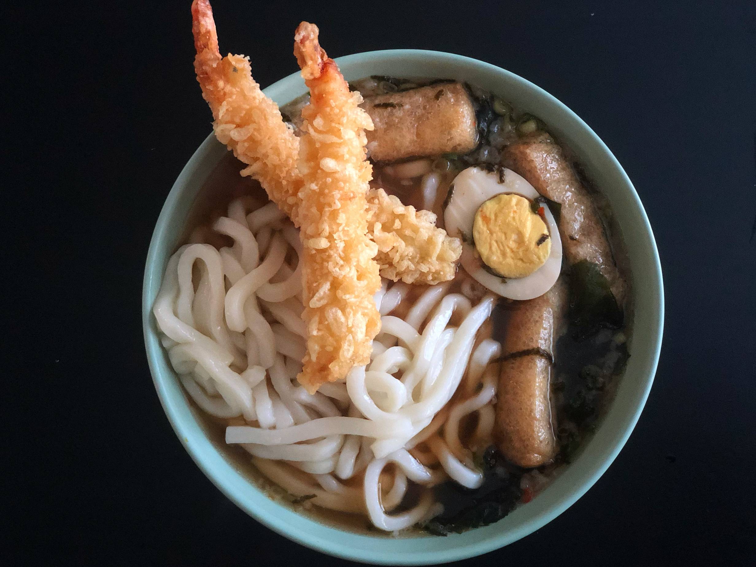 Two pieces of shrimp tempura rest on the side of the bowl and in the ramen. There are many udon noodles, some garnishes and a dark broth in a mint bowl on a black table. Photo by Alyssa Buckley.