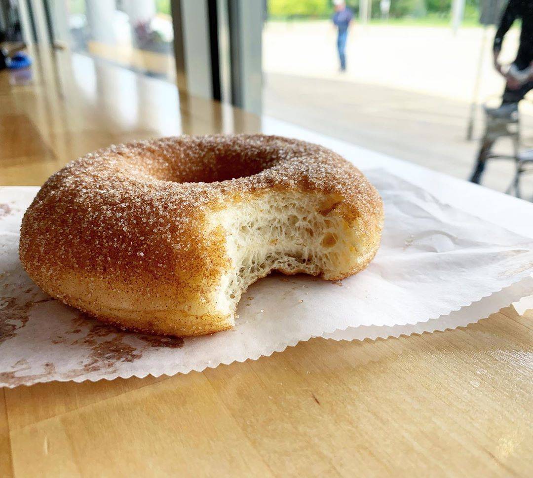 A cinnamon sugar doughnut sits with a bite out of it on a parchment paper sleeve. Image courtesy of Pandamonium Doughnuts Instagram.