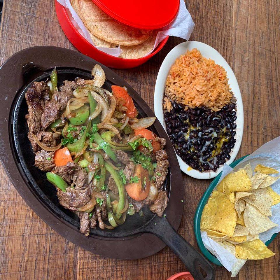 Steak fajitas sit in a cast iron pan on a wooden table next to corn tortillas and a side of rice and beans. Photo from Fiesta Cafe's Facebook page.