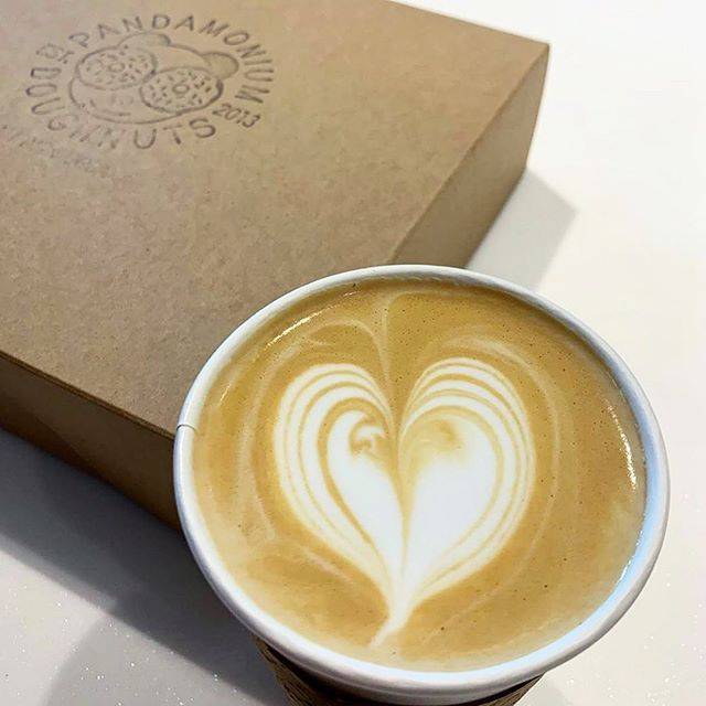 A light brown coffee drink with a heart swirl on top sits next to a closed paper box of Pandamonium Doughnuts. Image from Pandamonium Doughnuts Instagram.
