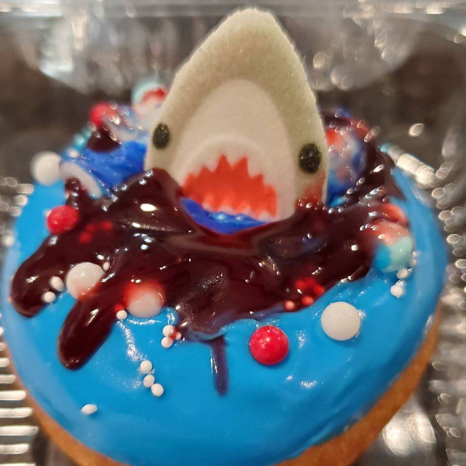 A donut with blue frosting, red icing, sprinkles, and a shark on top sits in a clear, plastic carryout container. Photo from Industrial Donut's Facebook page.