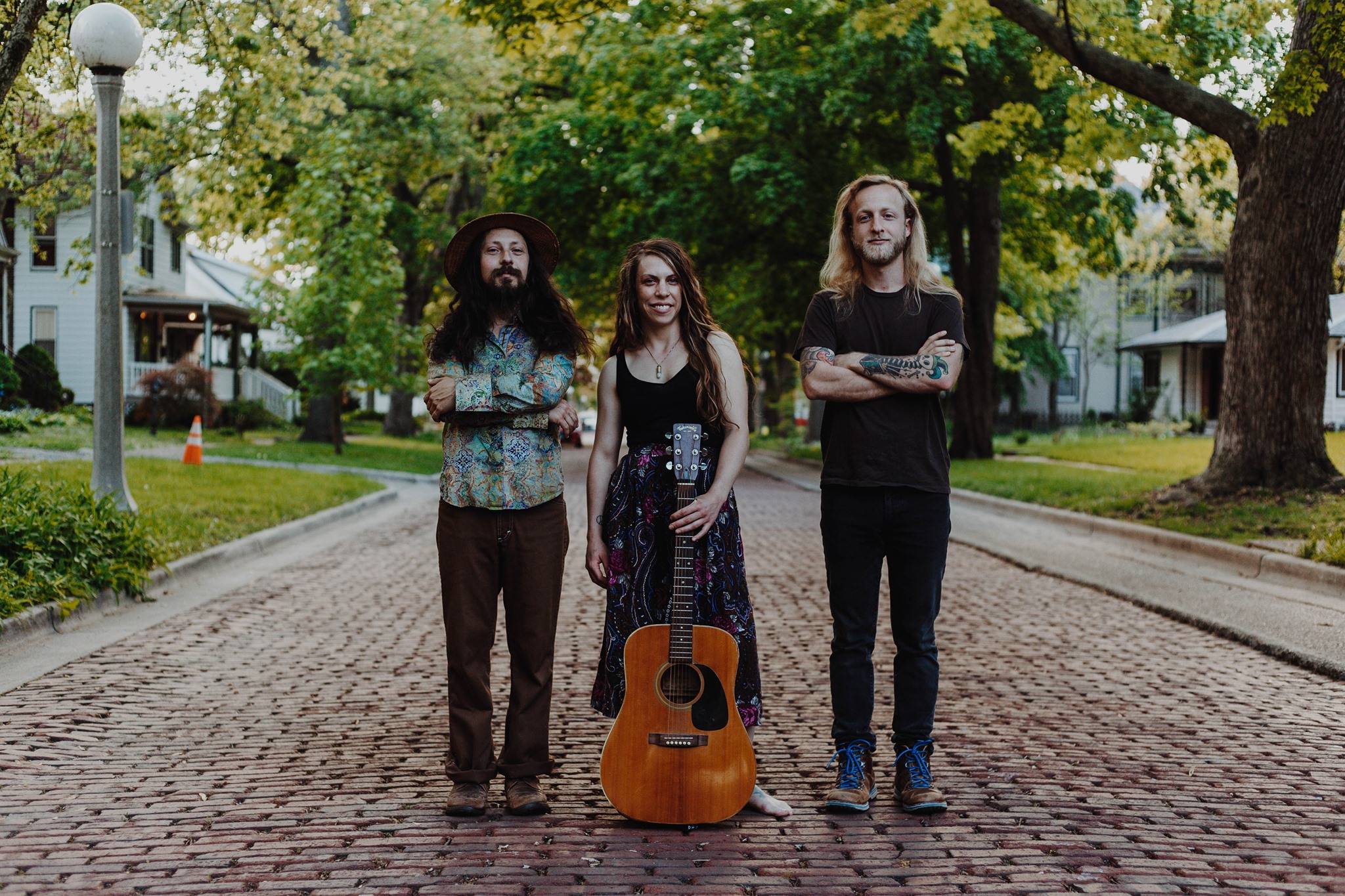 Three people stand in the middle of a brick-paved road. The woman in the middle is holding a guitar by her feet. Image by Veronica Mullen.