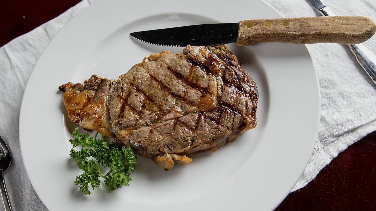 A 14 oz. bone-in ribeye with diamond grill marks rests on a white plate, which in turn sits on a small white cloth atop a wooden table. A wood-handled, serrated steak knife lays across the top of the plate, and a sprig of fresh parsley is tucked under the steak as garnish. Photo by Hamilton Walkerâ€™s.