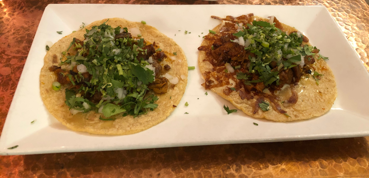A traditional and a Mexica taco al pastor from Maize. The tacos sit side-by-side on a white rectangular plate. Tacos are topped with onion and cilantro, and the Mexica taco has grilled cheese on the tortilla. Photo by Jesus Barajas.
