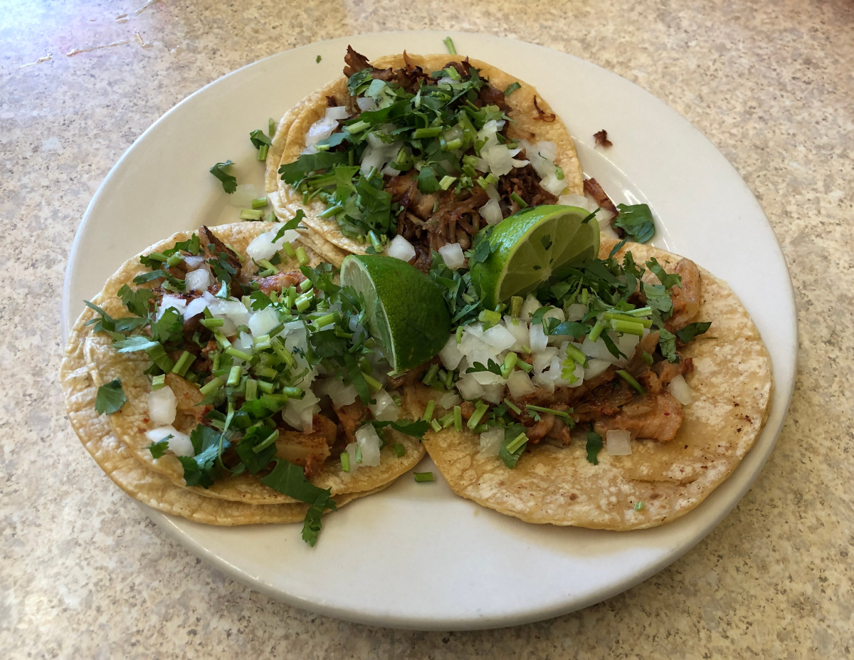 Two tacos al pastor and one carnitas taco from Mo's Burritos. The tacos are arranged in a triangular fashion on a circular plate. The carnitas taco is on top while the two tacos al pastor are on the bottom. Photo by Jesus Barajas.