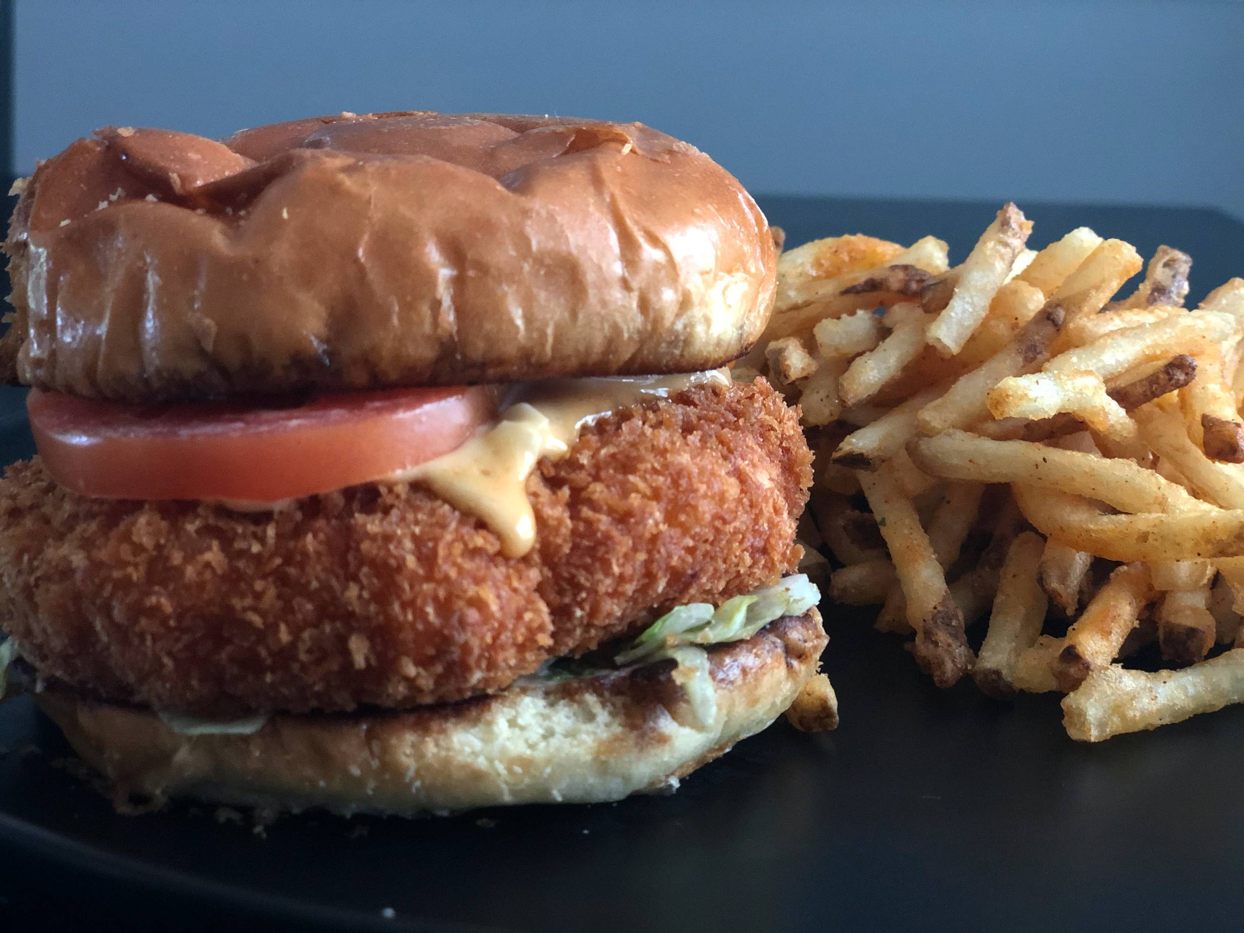 A burger with a crispy shrimp patty is sandwiched between a soft brown bun with tomato slices showing and a bit of lettuce below the patty. The burger sits on a plate next to a large pile of shoestring french fries. Photo by Alyssa Buckley.