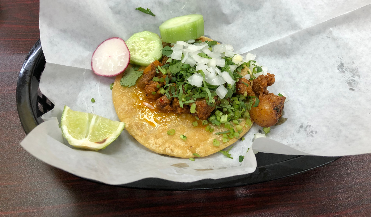 A taco al pastor from Tacos Locos. The taco is on parchment paper in a black plastic basket. The taco is garnished with cucumbers, a radish, and two lime wedges. Photo by Jesus Barajas.