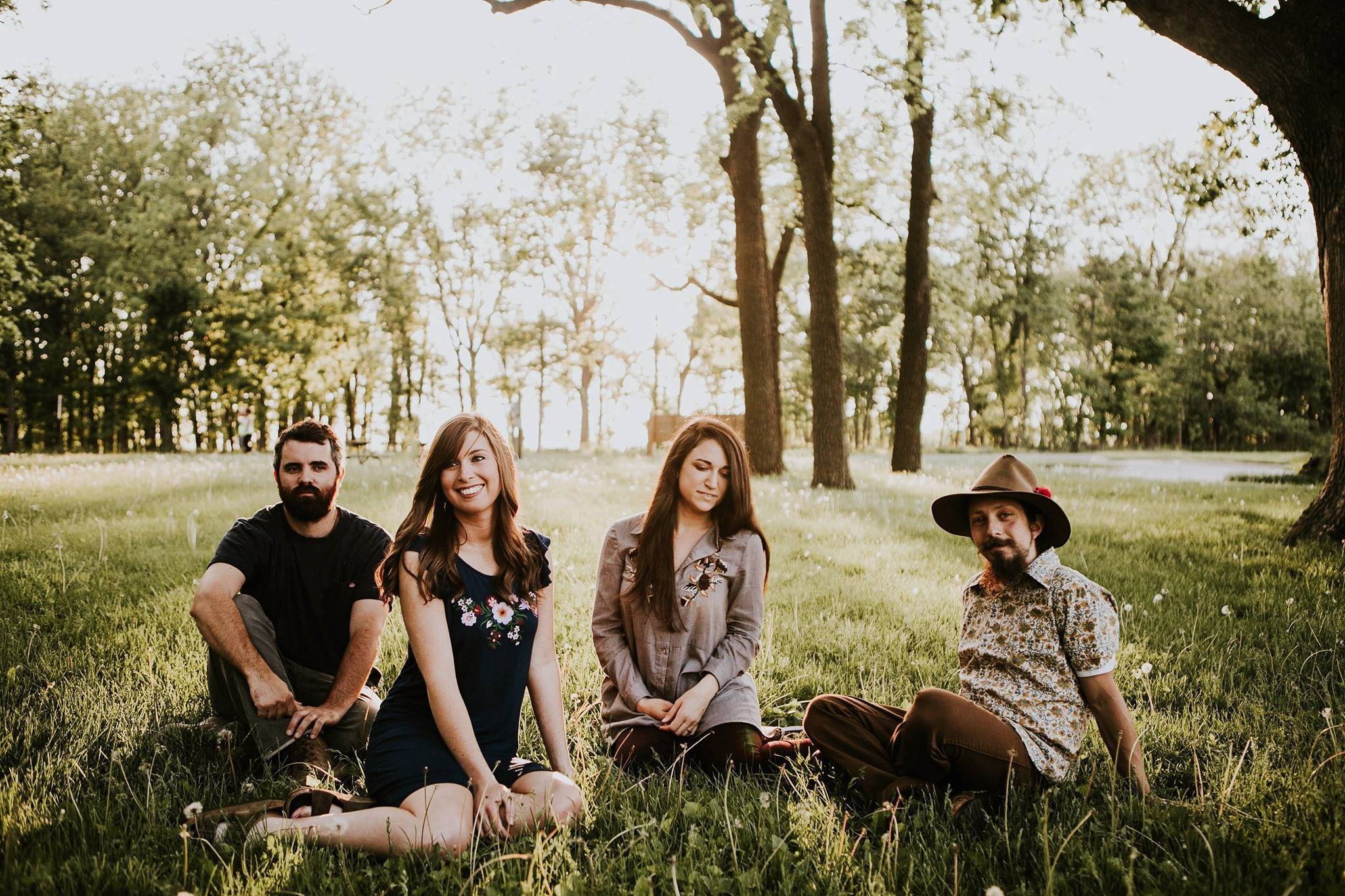 Four people sit in the grass, trees and wooded area behind them. One man sits on the far left, another man sits on the far right wearing a hat. Two women with brown hair sit in the middle. Photo by Anna Longworth. 