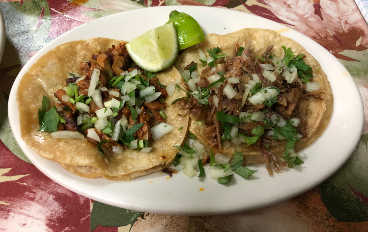 One taco al pastor and one carnitas taco from Huaraches Moroleon. The tacos sit side by side on a circular plate. They are garnished with onions, cilantro, and two lime wedges. Photo by Jesus Barajas.
