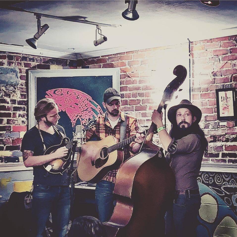 Three band members performing, one playing a mandolin, another playing guitar, another playing upright bass. There's a brick wall in the background. Photo from Hobnob String Band's Facebook page.