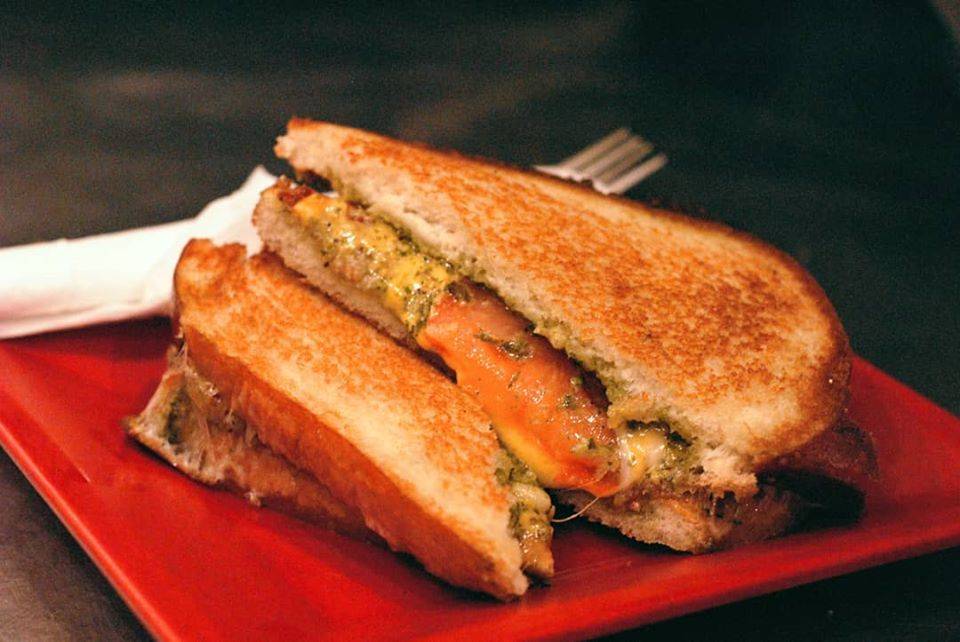 A diagonally sliced pesto grilled cheese sandwich from Aroma Cafe sits balanced on top of each other on a red plate with a white paper napkin rolled utensils. Photo from Aroma Cafe's Facebook page.
