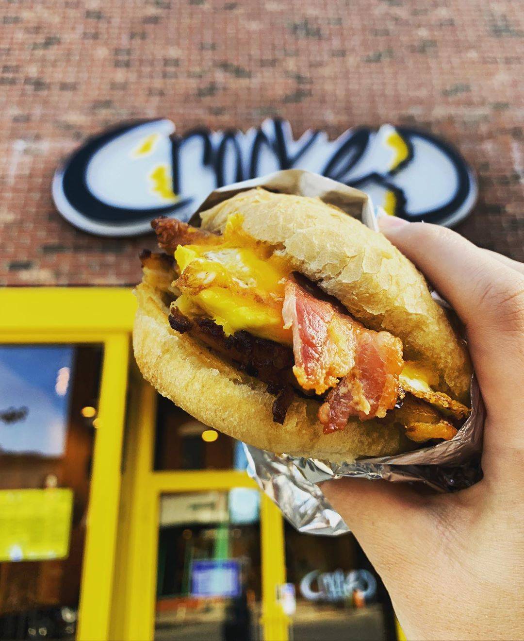A breakfast sandwich with bacon, cheese, and egg is in focus with the background of Cracked's Green Street location in the background. Photo provided by Cracked.