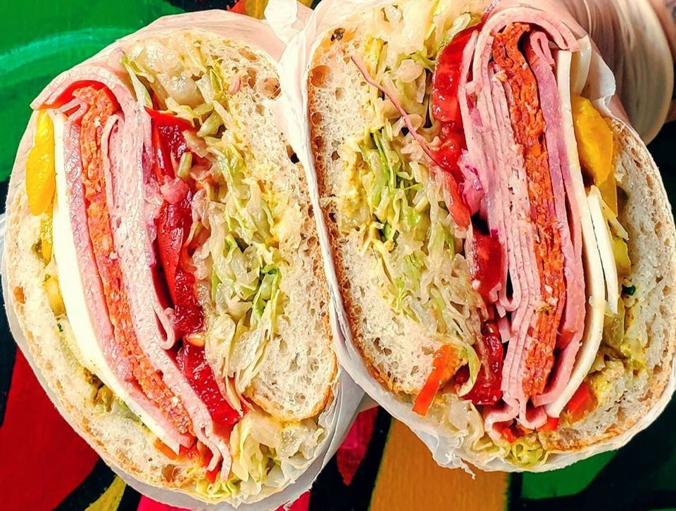 A sliced Italian sandwich is held, so both of the inside cuts are shown. The white ciabatta bread holds mortadella, pepperoni, ham, onions, lettuce, tomato, and peppers. Photo from Baldarotta's Facebook page.