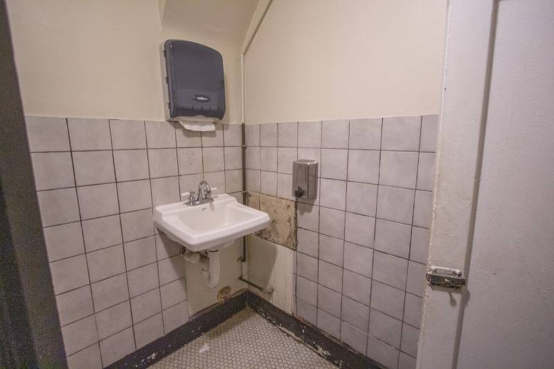 A bathroom sink hanging on a wall with white square tiles. A black paper towel holder is on the wall above it. A metal soap dispenser is on the adjacent wall. Some of the tiles are broken. Photo by Matt Wiley.
