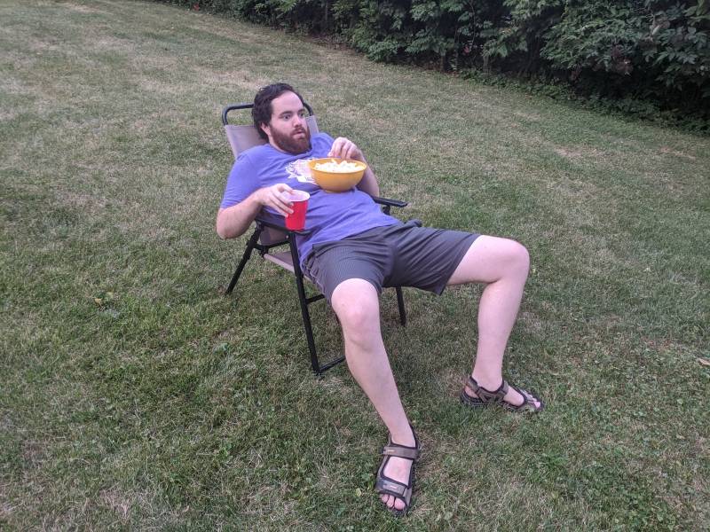 The writer is lounging in a lawn chair in the middle of a grassy lawn. He has a bowl of popcorn on his chest and is holding a red solo cup. Photo by Andrea Black.