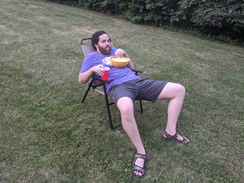 The writer is lounging in a lawn chair in the middle of a grassy lawn. He has a bowl of popcorn on his chest and is holding a red solo cup and has popcorn in his mouth. Photo by Andrea Black.