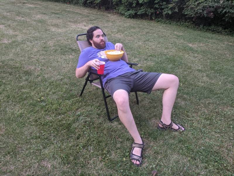The writer is lounging in a lawn chair in the middle of a grassy lawn. He has a bowl of popcorn on his chest and is holding a red solo cup. Photo by Andrea Black.