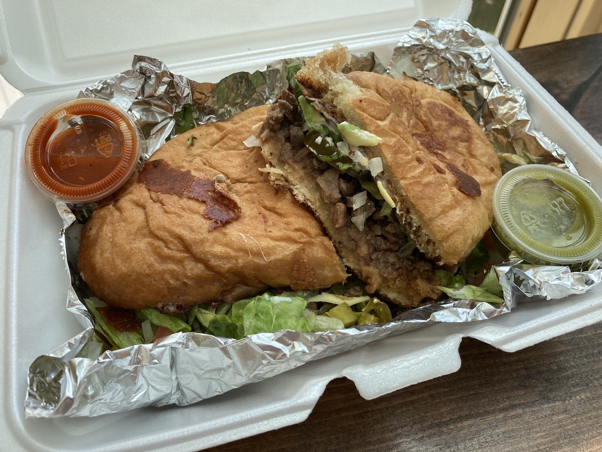 A steak torta sliced in half with a side cup of red salsa and a side cup of green salsa are in a tin-foiled lined white styrofoam takeout container. Photo by Anthony Erlinger.