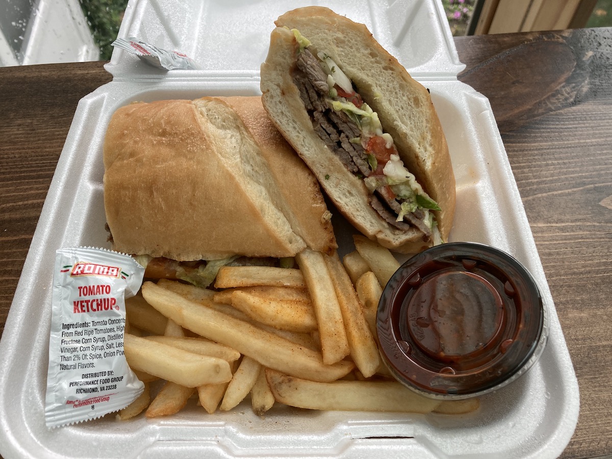 A steak torta on a French style baguette from Huaraches Moroleon sits in a white styrofoam container with a side of french fries, a packet of ketchup, and a small cup of red salsa. Photo by Anthony Erlinger.