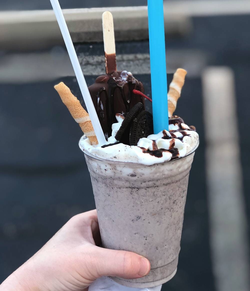 A Deluxe Oreo milkshake at El Oasis in Urbana, Illinois. A large, clear, plastic cup contains an Oreo milkshake and is garnished with whipped cream, cookies, an ice cream pop. A blue straw and a long plastic spoon are stuck in the milkshake. A white person's hand holds the cup. Photo by Jessica Hammie. 