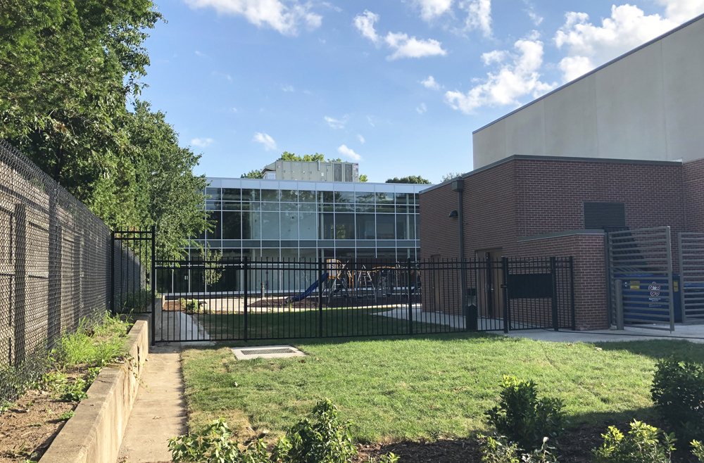 The backside of Dr. Howard elementary school in Champaign, pictured from West Park Avenue. There is a metal fence, beyond which is a courtyard with a playground. The building walls are made of glass, with a clear view into the two story building. Photo by Jessica Hammie. 