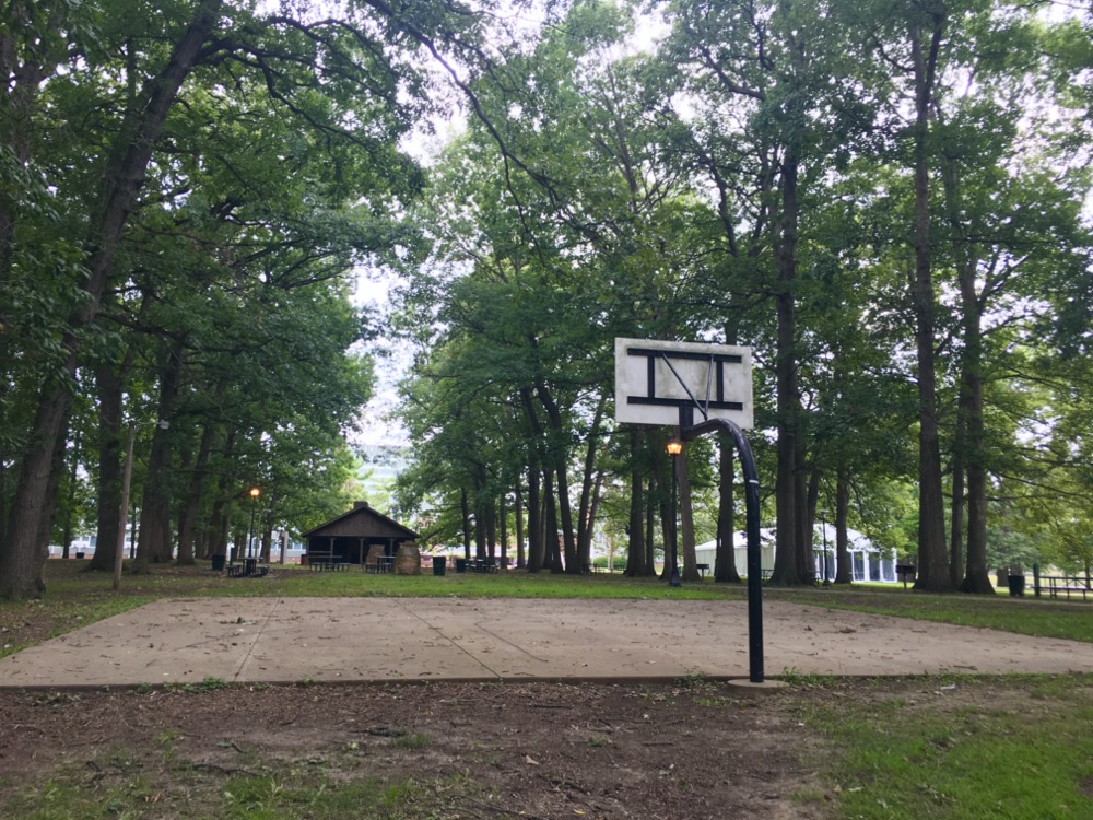 A basketball court, viewed from behind the hoop. The court and hoop are surrounded by green trees and grass. Photo by Osiris Ramos.