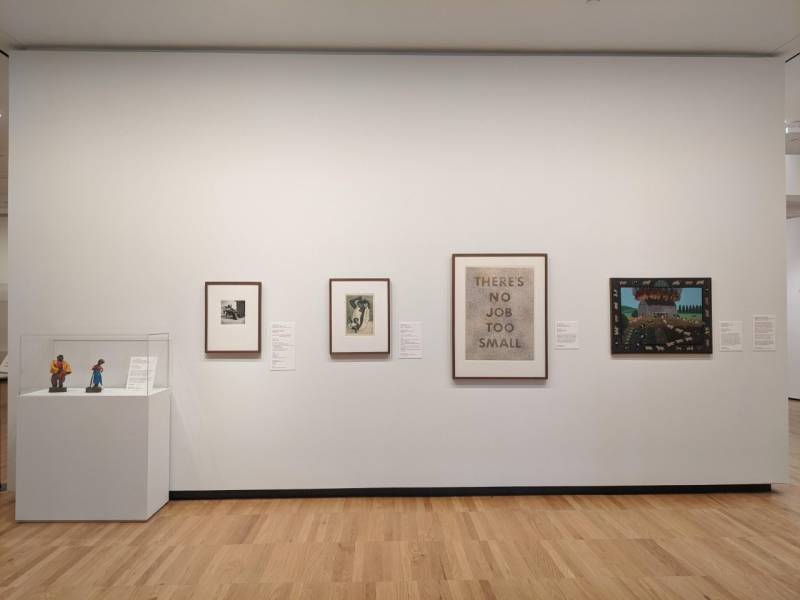 A white wall with four framed pieces of art hanging on it. There is a display case on the left with two figures in it. 