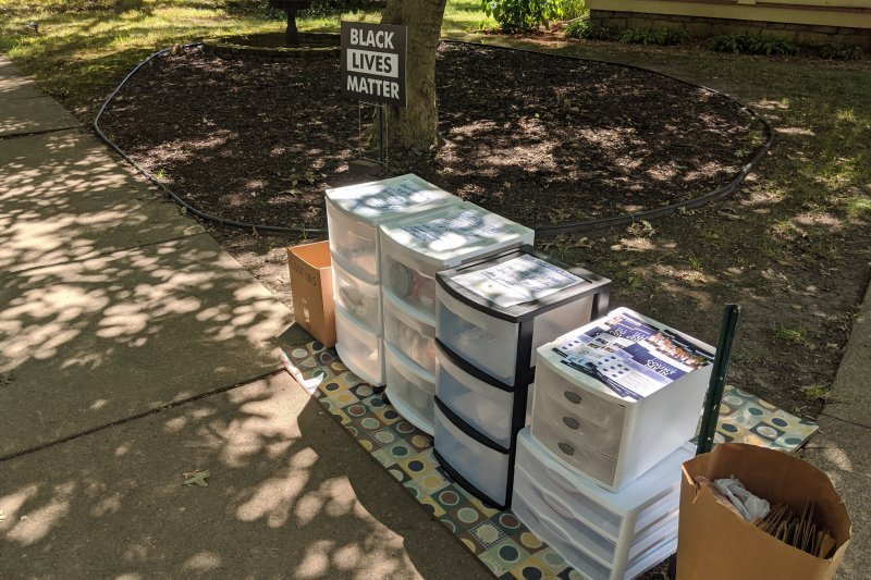Five sets of plastic drawers sit on a small area rug by a sidewalk. There is a paper bag full of other paper bags on one side of the drawers. A cardboard box is on the other side. A Black Lives Matter sign is in the yard behind the boxes. Photo by Andrew Adams.