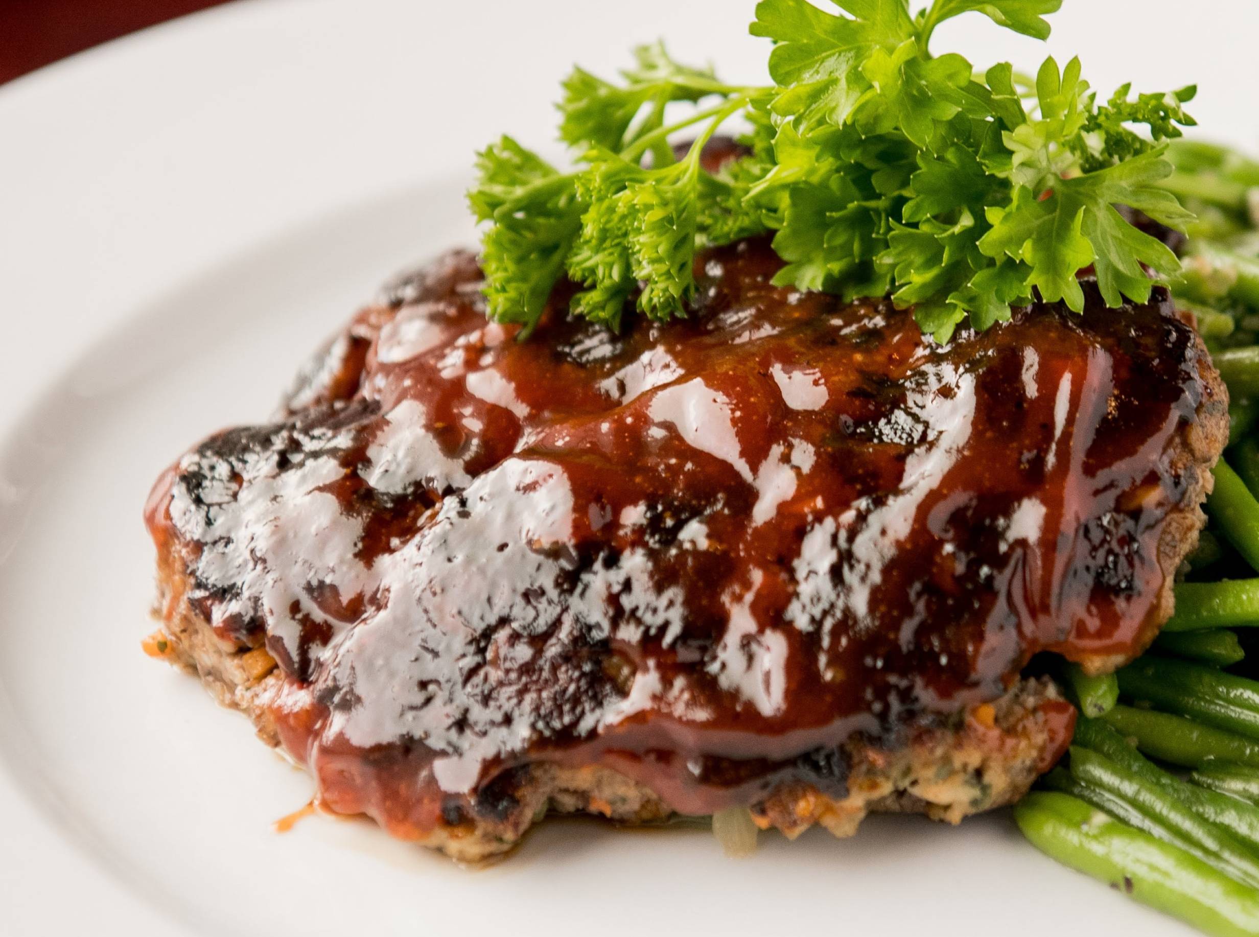 A single-serving, made-to-order meatloaf is placed on a round, white plate. A layer of zesty sauce and a sprig of fresh parsley top the meatloaf, which itself partially sits on a bed of fresh garlic green beans, cooked tender crisp, and roasted red potatoes. Photo by Hamilton Walkerâ€™s. 