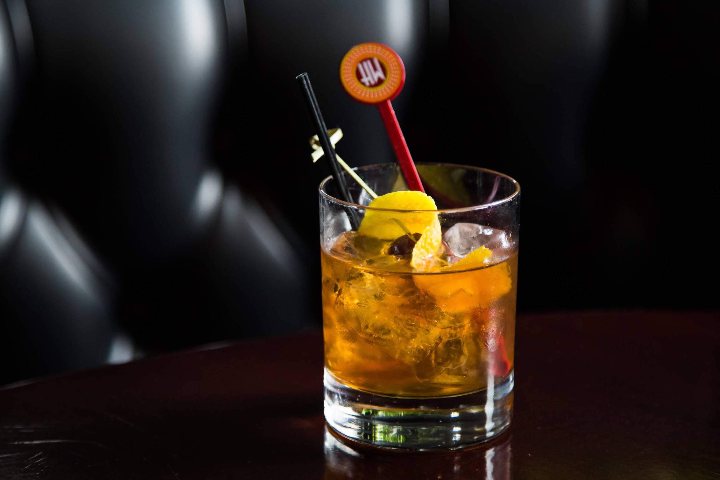 A thick-bottomed rocks glass sits on a dark wood table. In the background is the diamond tufted pattern of the black leather booth at which the drink is served. The drink is Â¾ full of large, clear ice cubes and a bourbon cocktail. A red Hamilton Walkerâ€™s swizzle stick and an orange peel twisted around a cherry, both speared on a bamboo toothpick, accent the drink. Photo by Hamilton Walkerâ€™s.