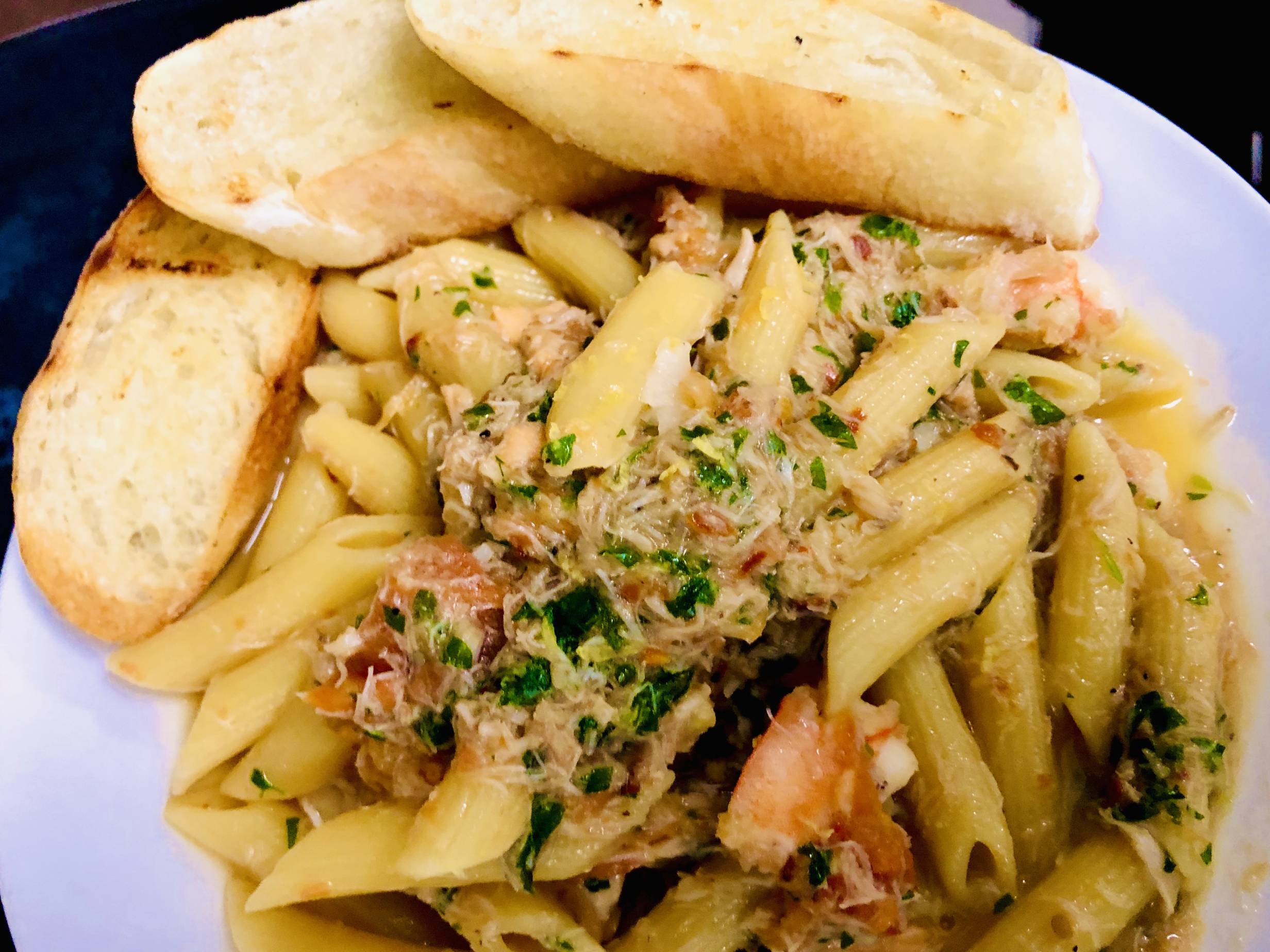 Penne pasta is topped with a slightly golden, glossy spiced tomato butter sauce, which pools at the bottom of the dish white pasta bowl. A combination of flaked crab, salmon, and a few whole shrimp are scattered throughout. It is garnished with parsley flakes. Three thin, bias-cut slices of lightly grilled baguette are laid, overlapping, across the top of the dish. Photo by Hamilton Walkerâ€™s.