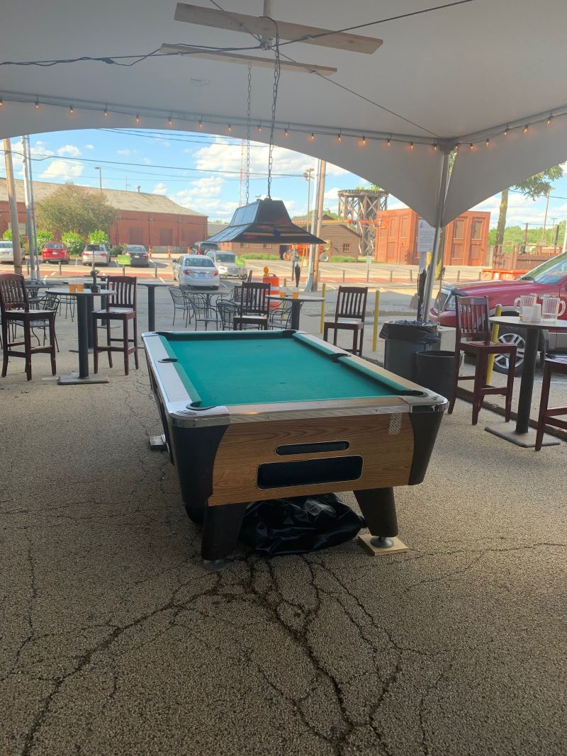 A pool table with green felt sits in a parking lot under a large white tent. There are high top bar tables along the outskirts of the tent. Photo by Seth Fein.