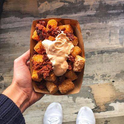 A paper basket of fried tots covered in cheese and bacon is held by a white hand over a gray wooden floor. Photo provided by Cracked.
