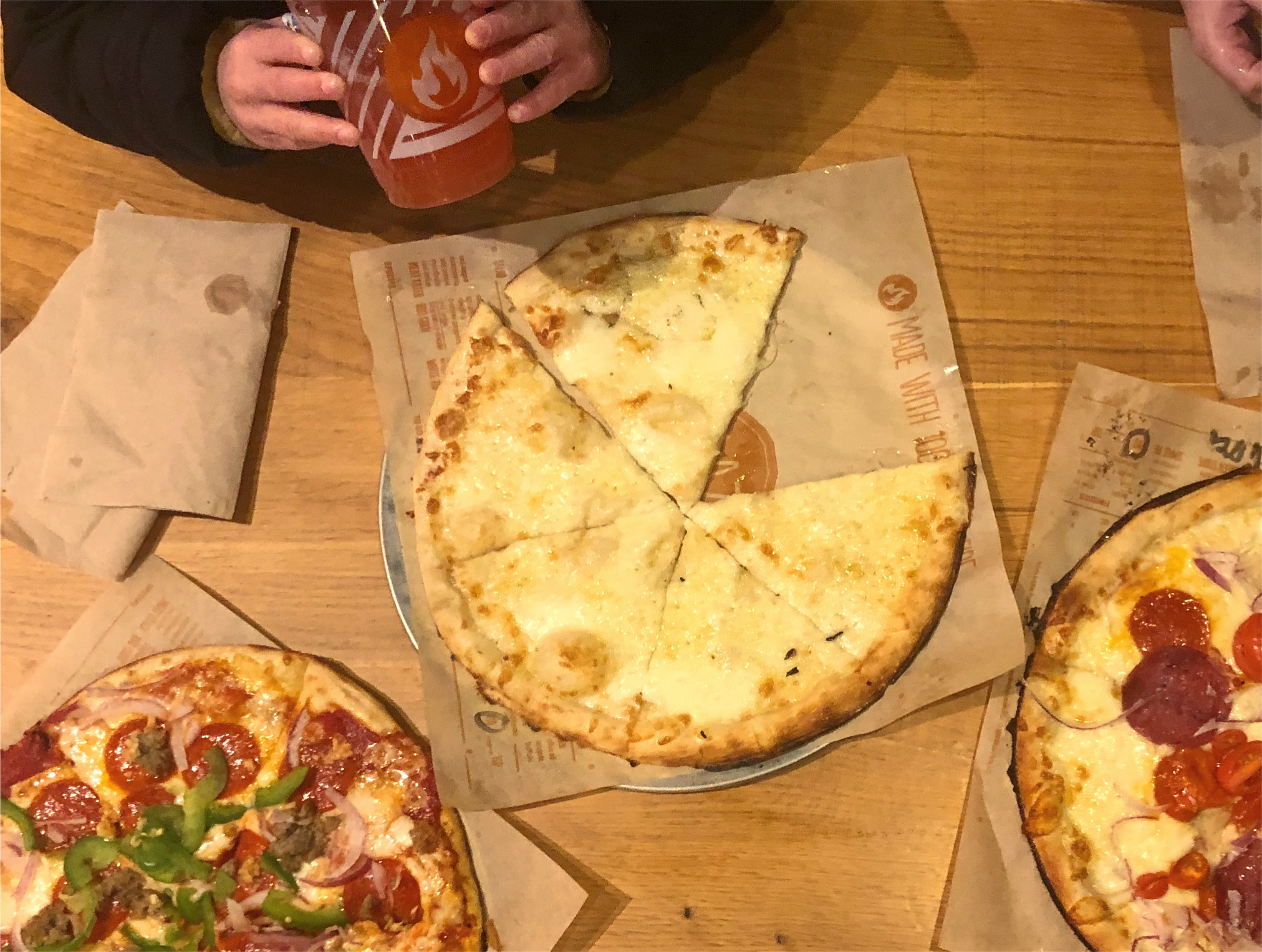 An overhead shot of a wooden table at Blaze Pizza. Two pizzas with toppings are cut off on the bottom, and in the center is a plain cheese pizza with one slice missing. Photo by Alyssa Buckley.