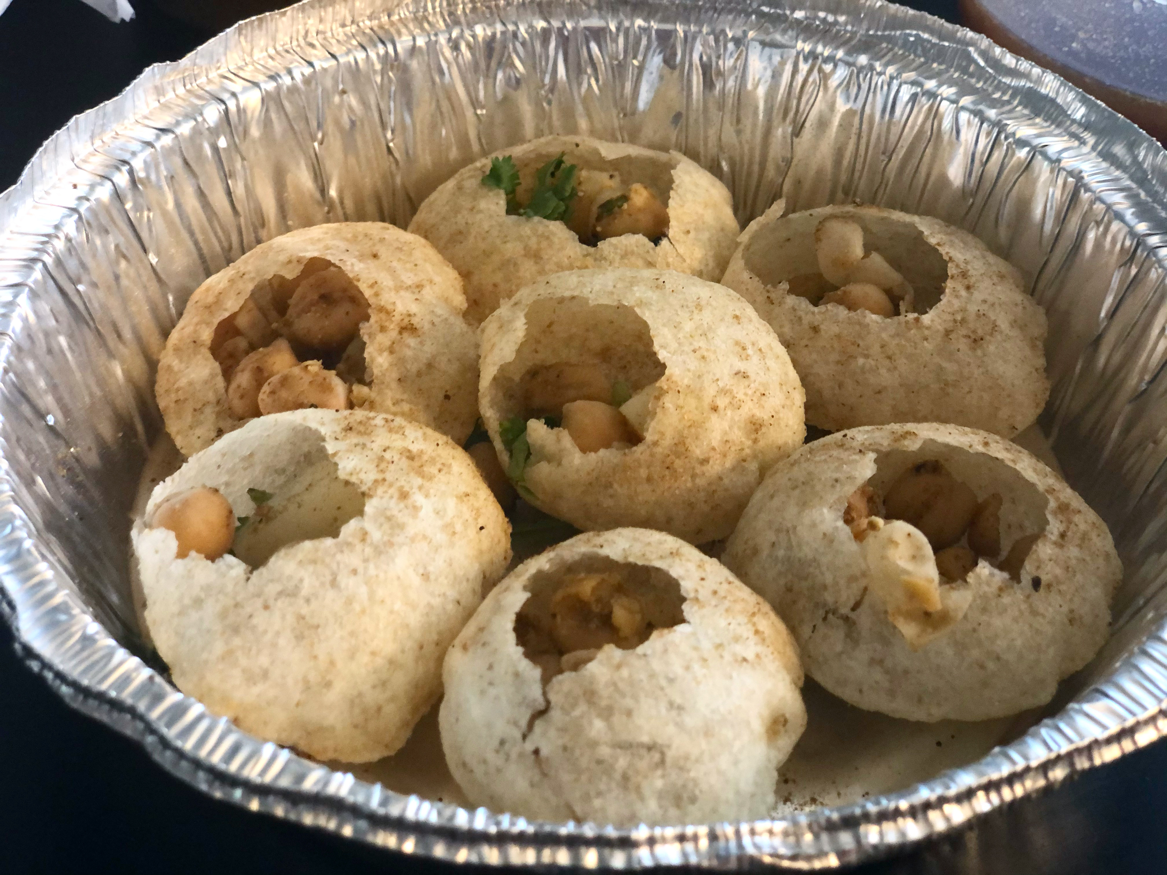 Seven small spheres are cracked open at the top revealing chickpeas and cilantro in a circular tin carryout container. Photo by Alyssa Buckley.