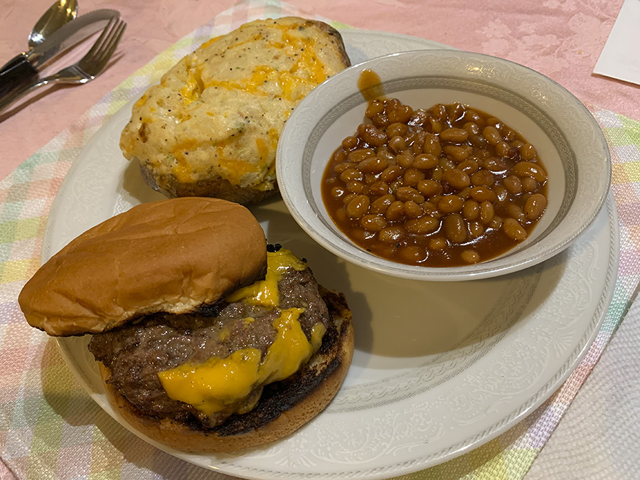 A photo of the author's dinner: cheeseburger with twice baked potato and baked beans on a paper plate on a pink table. Photo by Zoe Valentine.