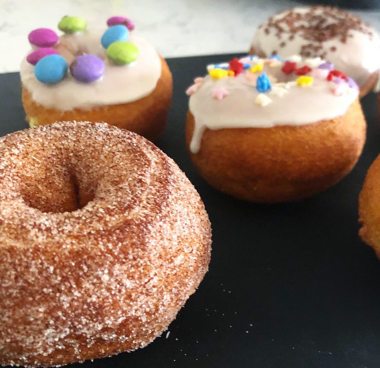 Donuts with varying toppings are on a black cutting board. The closest one is tossed in cinnamon sugar, and the others are frosted with white icing. Photo by Alyssa Buckley.