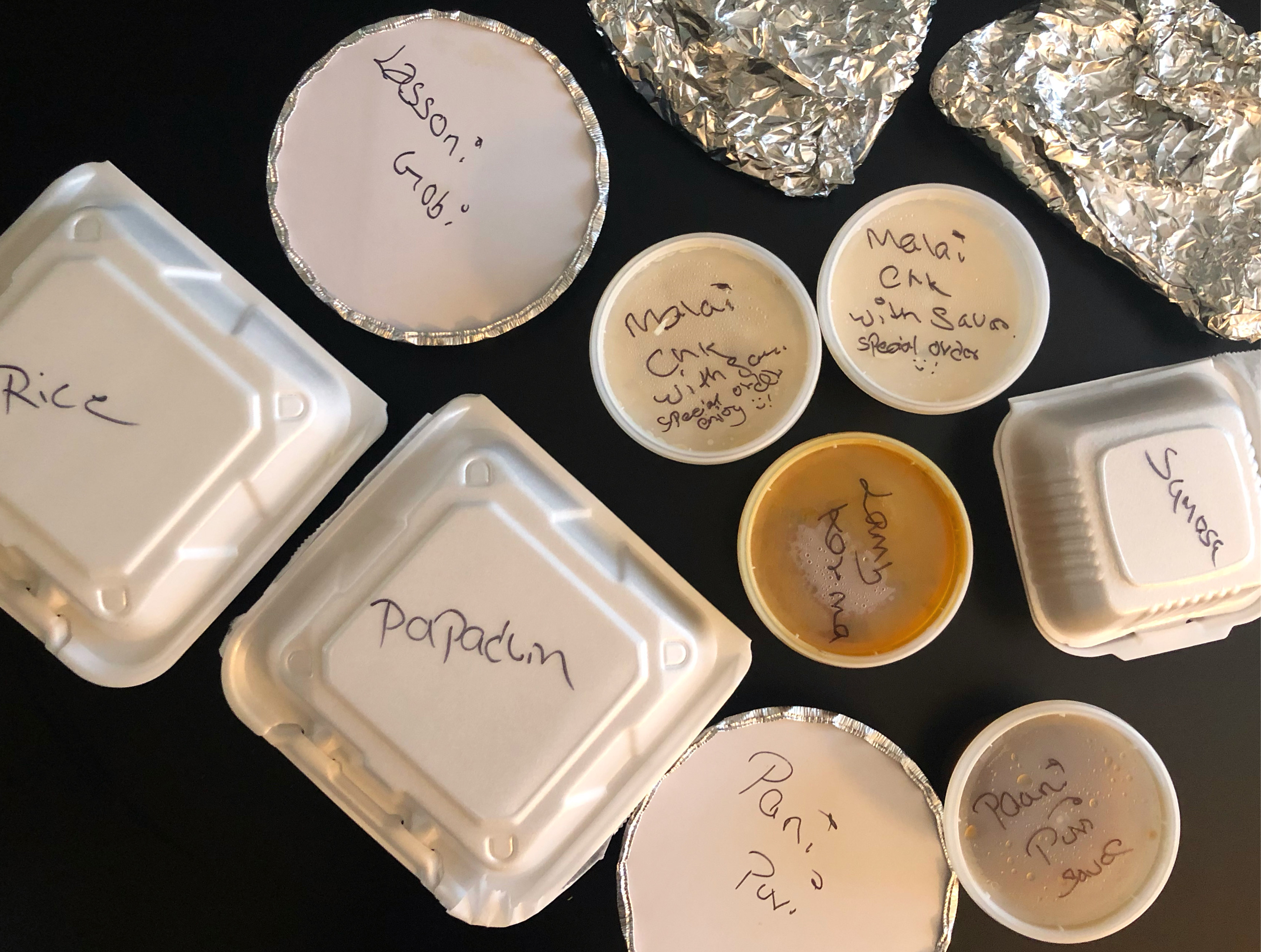 A collection of takeout containers from Himalayan Chimney are scrawled in black ink the names of the dishes on a black table. Photo by Alyssa Buckley.